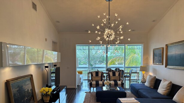 A modern living room with a sofa, a coffee table, and a TV. There are large windows and a chandelier. - Spacious 4-bedroom apartment At IMG Academy (Bradenton)