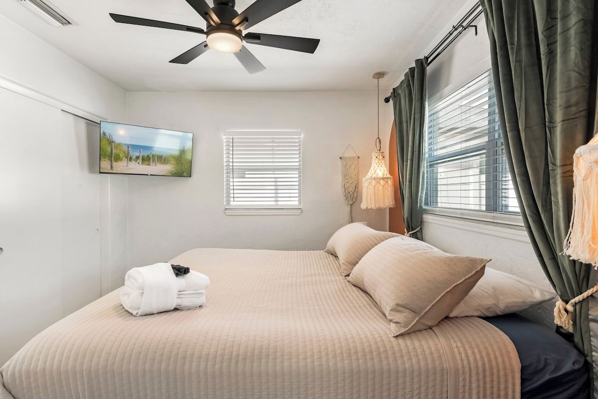 A hotel room with a bed, a ceiling fan, a television, a window with blinds, and a wall-mounted picture.