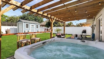 A hot tub under a wooden pergola with a clear view of a backyard, including a barbecue area and a small shed.