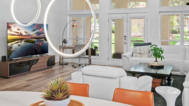 Een moderne woonkamer met een flatscreen televisie, witte banken, een eettafel met een plant als middelpunt en grote ramen.