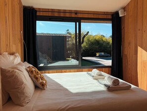 Ein Schlafzimmer mit großem Fenster, einem Bett mit weißer Bettwäsche, einem zusammengerollten Handtuch und einem Kopfkissen mit braunem Muster.