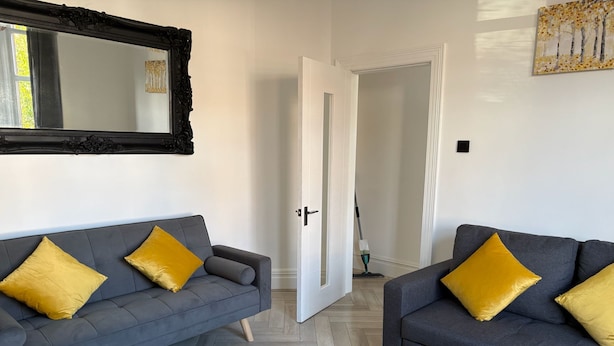 A modern living room with two grey sofas, each with a single yellow pillow, a large mirror with an ornate frame, and a wooden herringbone floor. - Coastal Retreat, Rhos-on-Sea, 1-Min Walk to Beach (Rhos on Sea)