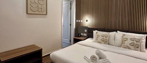 Un dormitorio moderno con una cama grande, una mesita de noche, un banco de madera, una lámpara de pared y una obra de arte enmarcada.