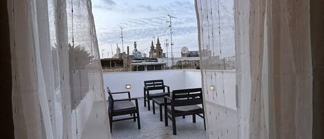 A view through sheer curtains to an outdoor seating area with tables and chairs, a building with a dome in the background, and a clear sky.