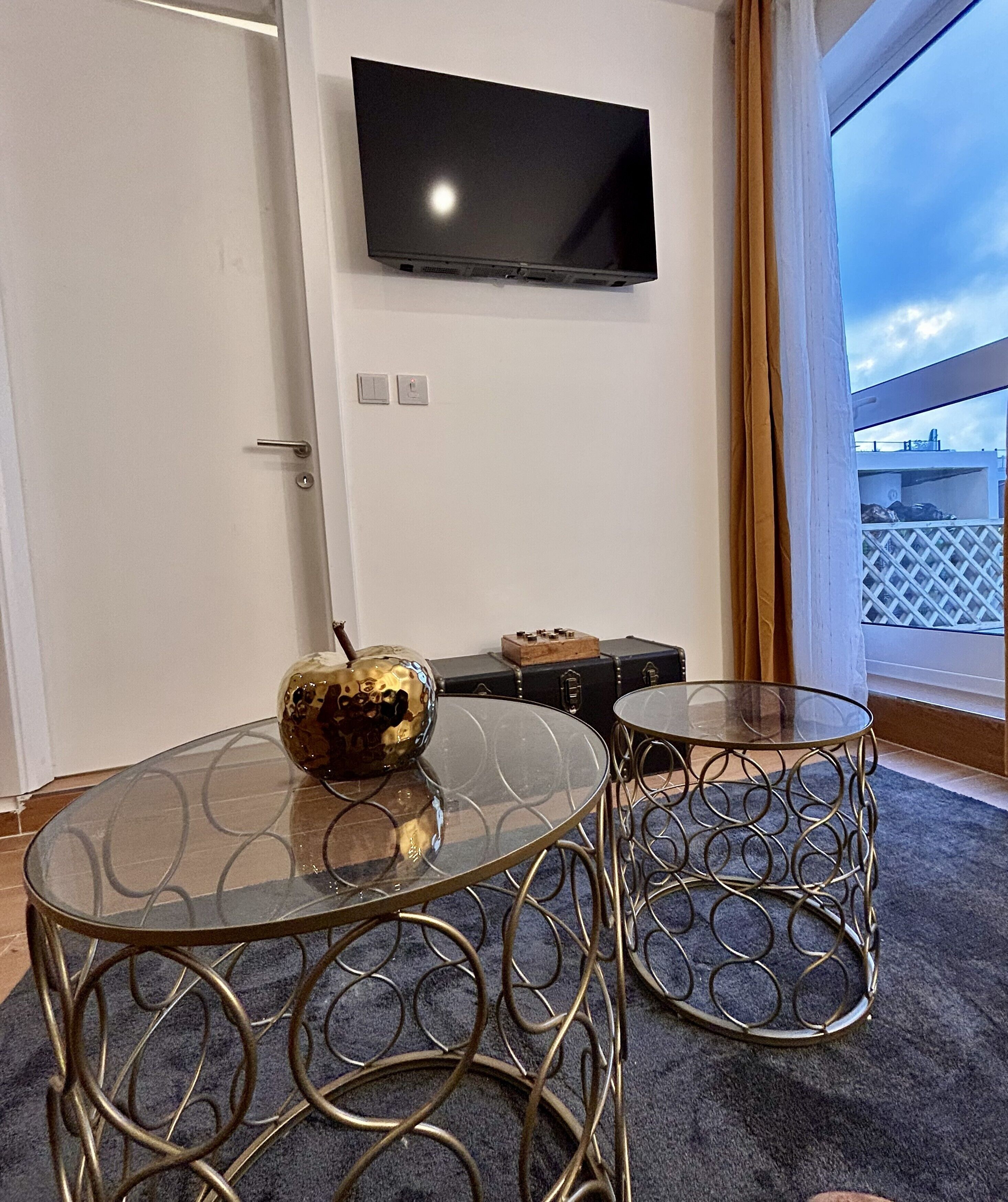 A room with a flat-screen TV mounted on the wall, two glass-top tables with metal frames, a decorative globe on one table, and a view of the outside through a window.