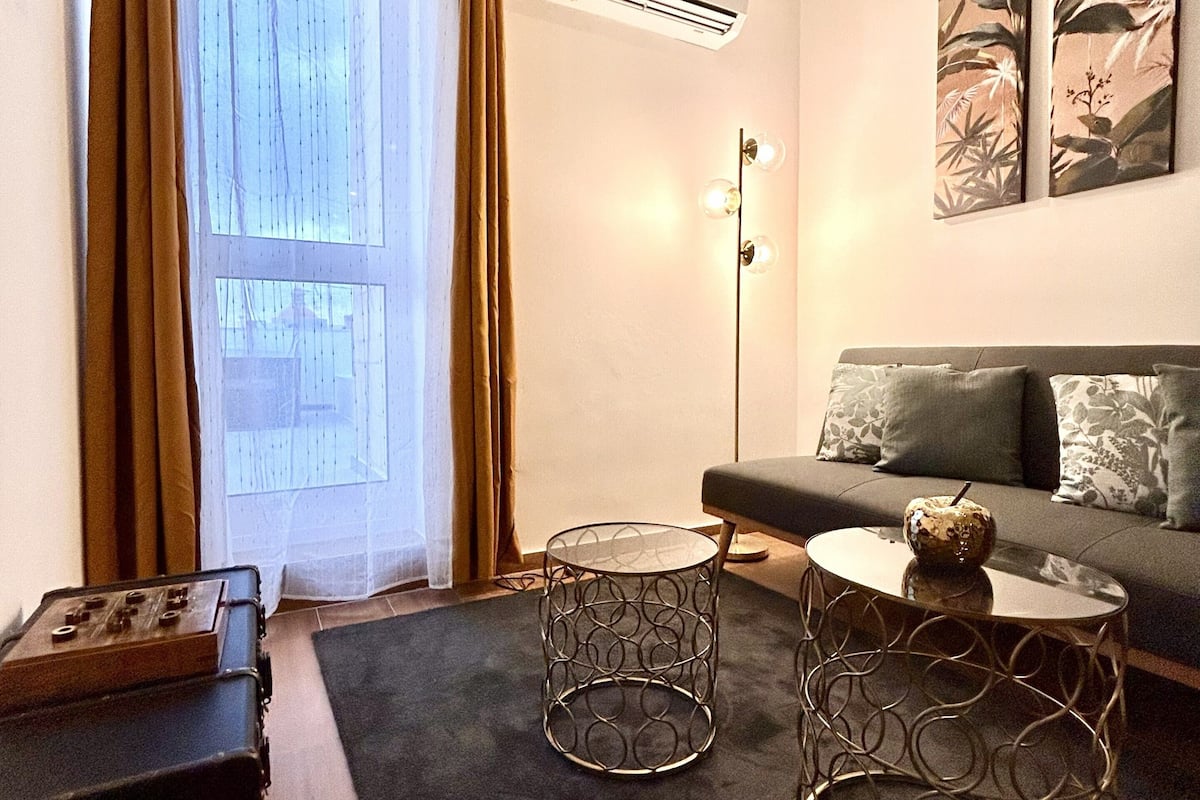 A living room with a sofa, two round tables, a suitcase, and a large window with curtains.