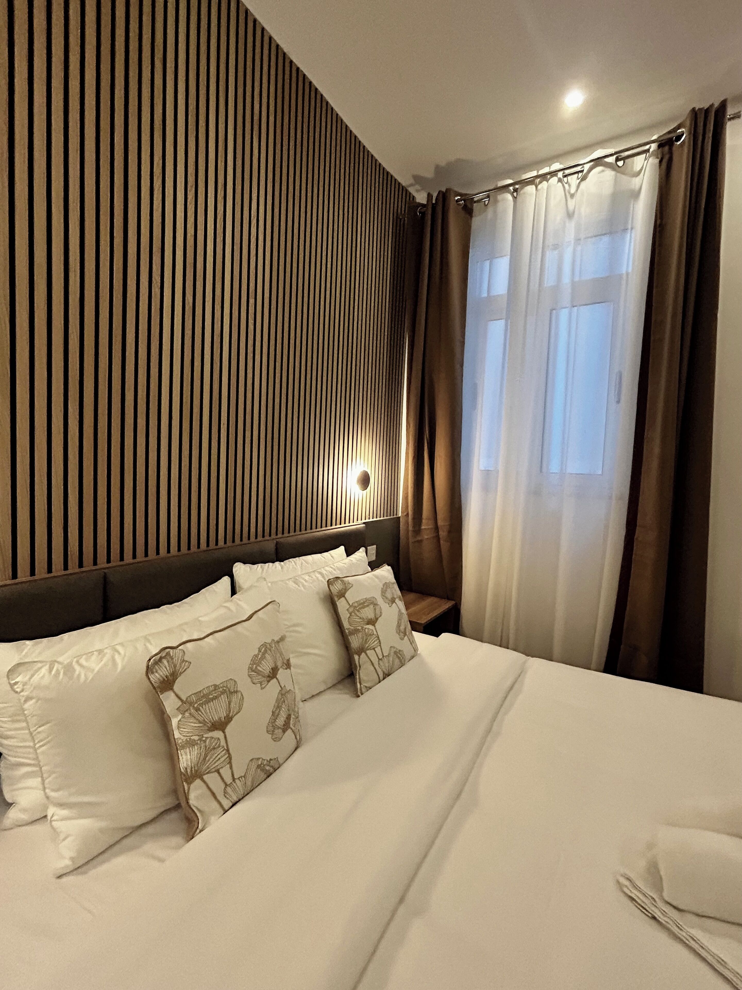 A hotel room with a bed, a headboard, a nightstand, and a window with curtains.