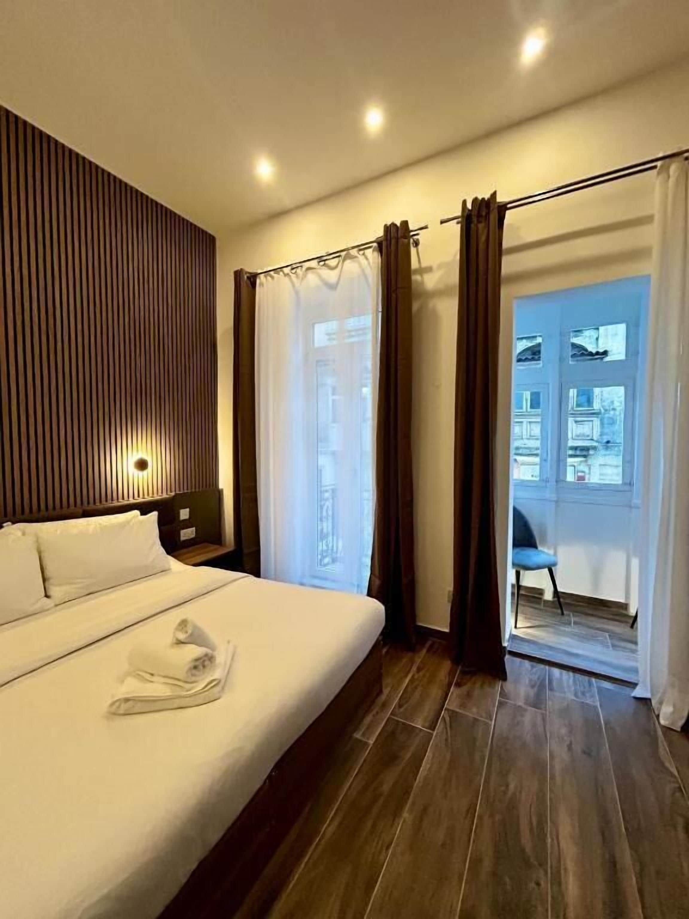 Une chambre d’hôtel moderne avec un grand lit, une tête de lit en bois, une table de chevet et une vue sur le balcon extérieur.