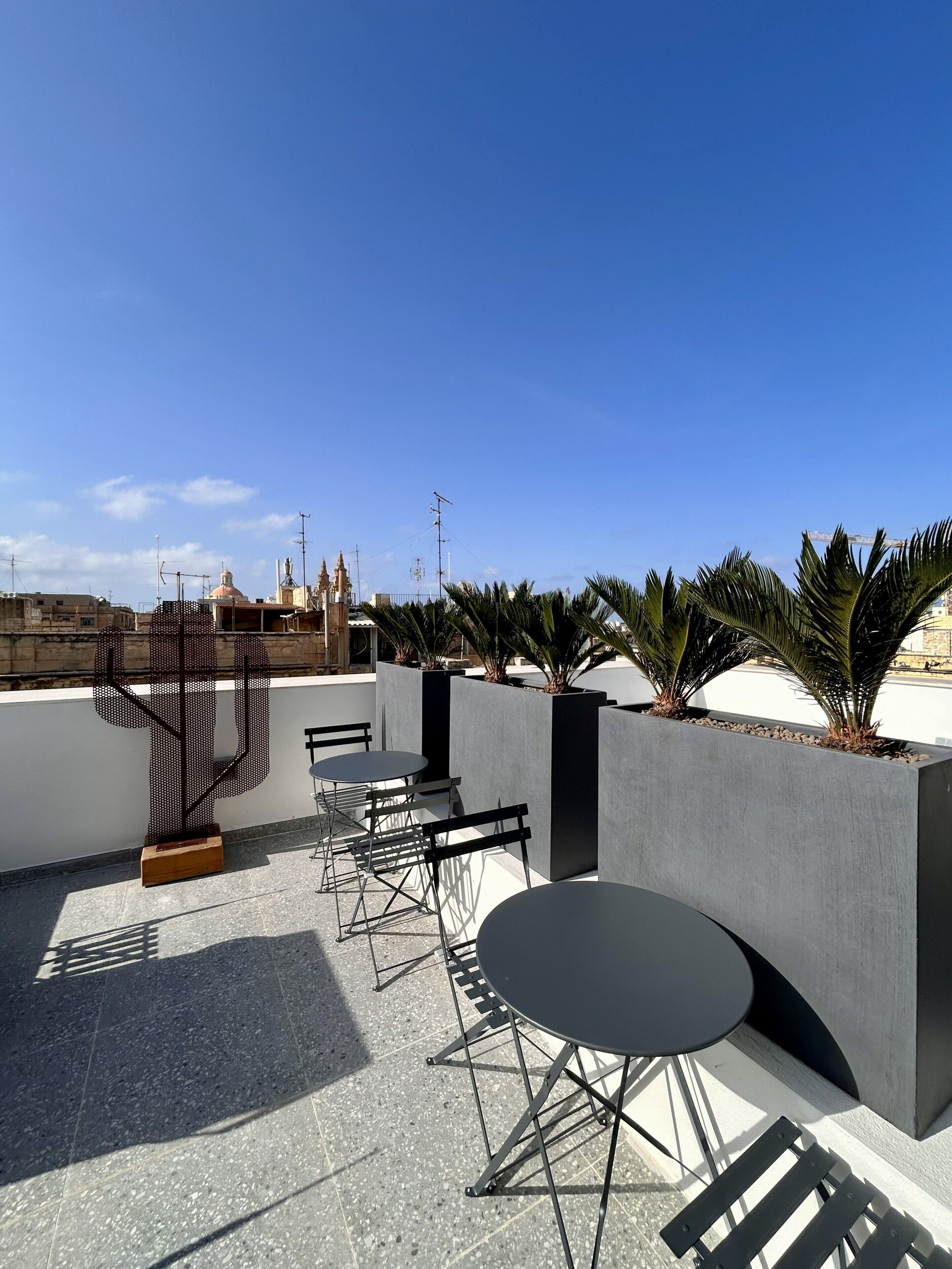 An outdoor seating area with tables and chairs, a cactus sculpture, and a view of buildings in the background.