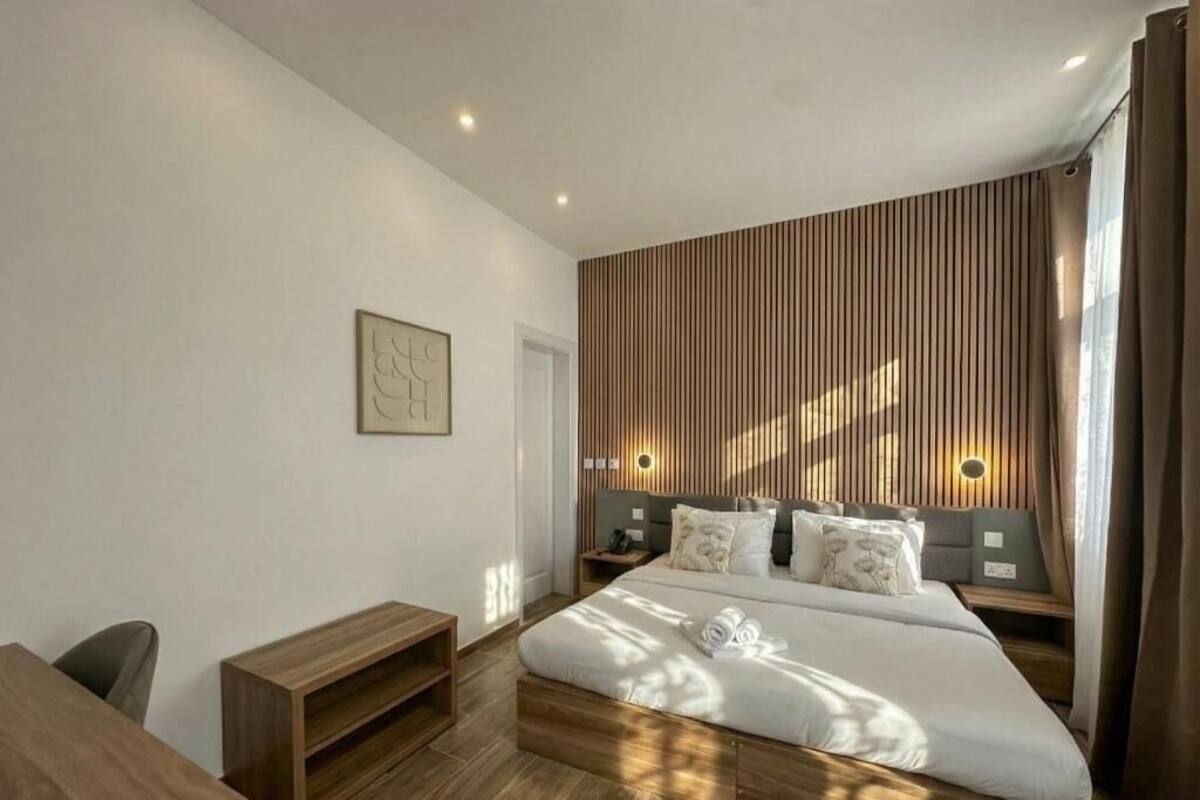A modern bedroom with a large bed, wooden headboard, bedside tables, and a wall-mounted light fixture.