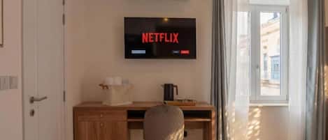 A room with a wooden desk, a chair, a television mounted on the wall displaying 'NETFLIX,' and a window with curtains.
