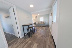 Ein Zimmer mit Holzboden, einer weißen Tür, einem kleinen Tisch mit Hocker, einem Sofa und einem Fenster mit Jalousien.