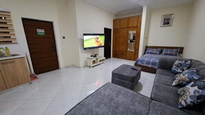 A living room with a sofa, ottoman, TV, wooden cabinet, and a door with a sign. - Savvy Studio Apt@RealShala Galaxy-School-Adjiringanor (Ashaley Botwe)