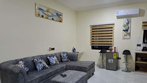 A living room with a grey sectional sofa, a small ottoman, a TV, a refrigerator, and a wall-mounted air conditioner. - Savvy Studio Apt@RealShala Galaxy-School-Adjiringanor (Ashaley Botwe)
