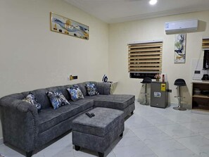 A living room with a grey sectional sofa, a small ottoman, a TV, a refrigerator, and a wall-mounted air conditioner. - Savvy Studio Apt@RealShala Galaxy-School-Adjiringanor (Ashaley Botwe)