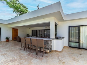 A modern outdoor kitchen with a stone island, bar stools, and a ceiling fan. - Connections Encuentro Tropical Oasis DR (Cabarete)