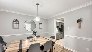 Un comedor con una mesa de madera, seis sillas, una lámpara colgante, dos espejos en la pared y una cocina visible a través de un vano abierto.