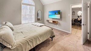 Un dormitorio con cama, televisor montado en la pared, ventana con persianas y puerta que da a un comedor.
