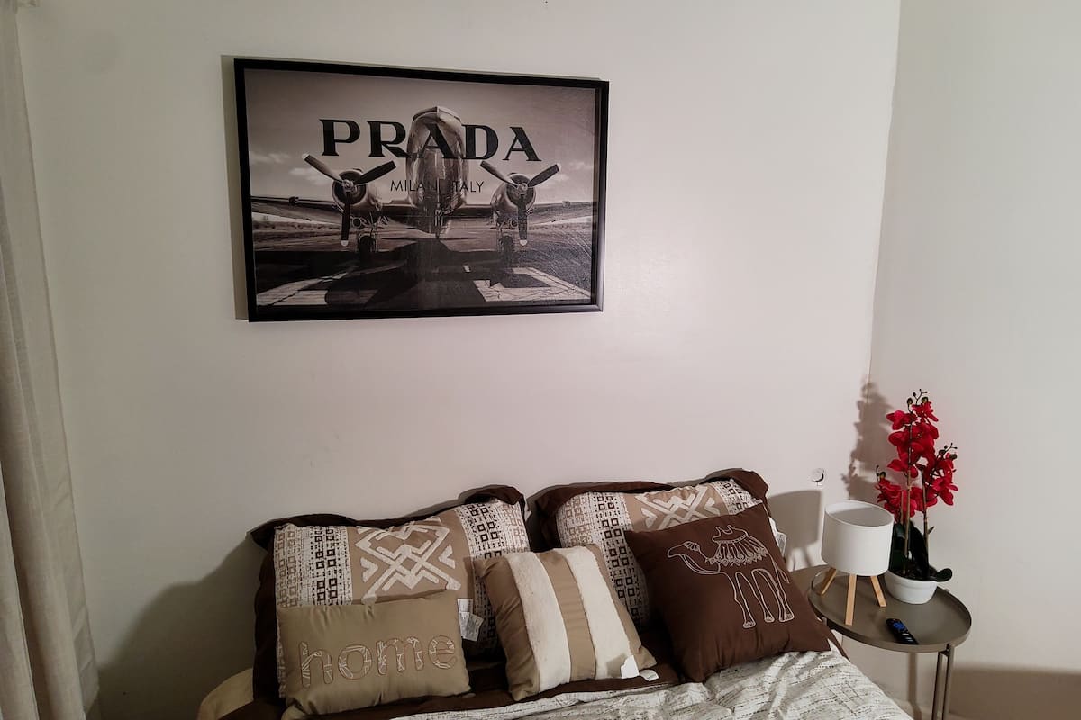 A bedroom with a bed, pillows, a framed picture of a car on the wall, a small table with a plant, and a remote control.