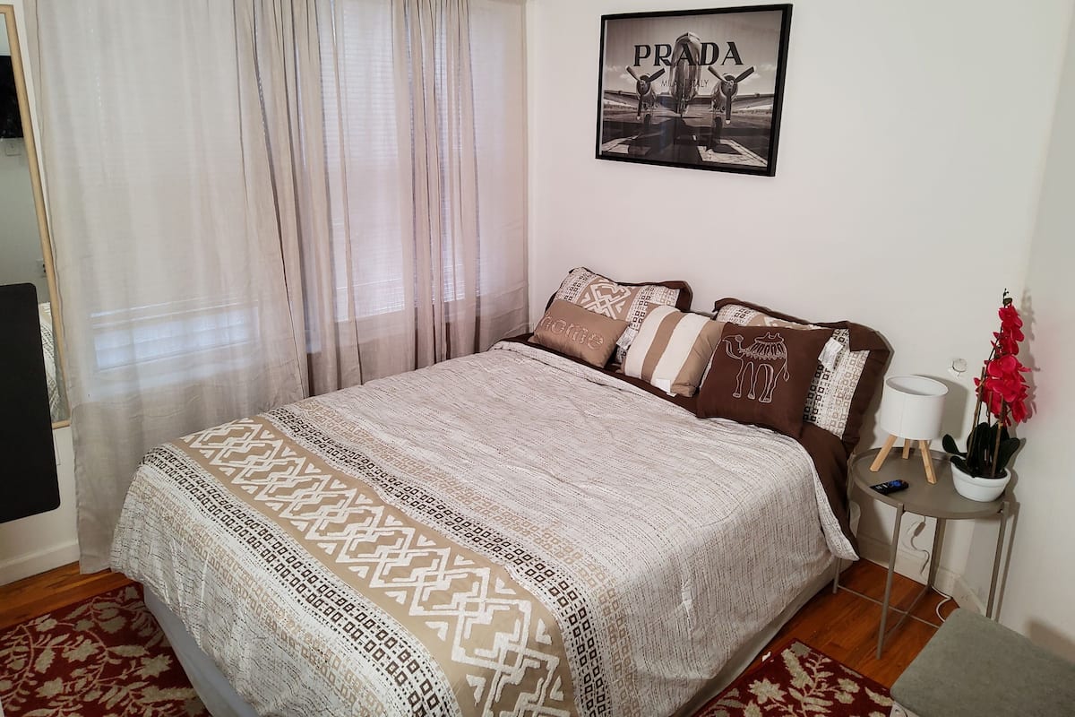 A bedroom with a bed, a framed picture on the wall, a bedside table with a lamp, and a red potted plant.
