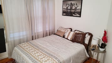A bedroom with a bed, a framed picture on the wall, a bedside table with a lamp, and a red potted plant.