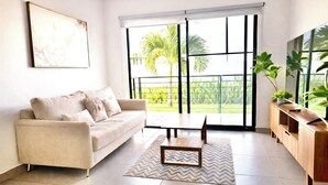 A living room with a sofa, a coffee table, a TV, and a view of a garden through large windows. - Resort-Style Condo- Pool, Patio, Playground + Palapa Lounge (Aldea Hortus)