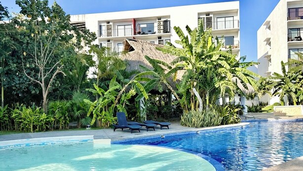 A swimming pool with lounge chairs, surrounded by tropical plants and a building with balconies. - Resort-Style Condo- Pool, Patio, Playground + Palapa Lounge (Aldea Hortus)