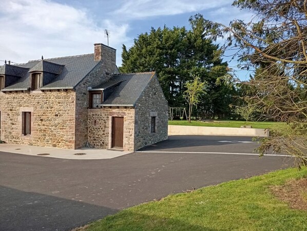 Un bâtiment en pierre avec un toit en pente, une allée et, au fond, un ensemble de jeux pour enfants avec une balançoire.