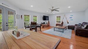 A spacious living room with a wooden dining table, a sofa, a TV mounted on the wall, and a ceiling fan. - Answered Prayers Main House-Step out to a peaceful morning or star gaze at night (Kingston)