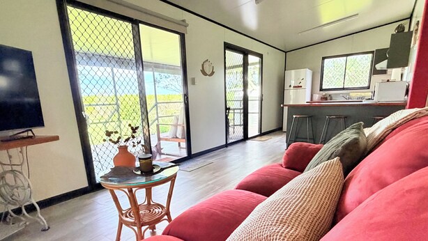 Ein Wohnzimmer mit einem roten Sofa, einem kleinen runden Tisch mit einer Vase, einem Fernseher auf einem Schrank sowie einer Küchenzeile mit Kühlschrank und Spüle.
