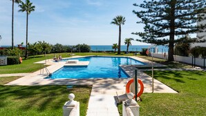 Piscina retangular com boia salva-vidas, rodeada por grama, palmeiras e vista para o mar.