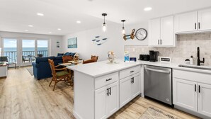 A modern kitchen with white cabinets, a built-in dishwasher, and a dining area with a blue sofa and wooden chairs. - Oceanfront 2BR Condo | Steps to Beach | Sleeps 8 (Ocean City)