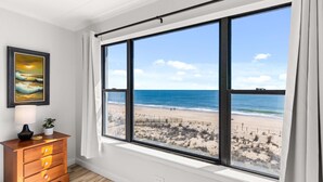 A room with a large window overlooking a beach, a wooden dresser, and a framed painting on the wall. - Oceanfront 2BR Condo | Steps to Beach | Sleeps 8 (Ocean City)