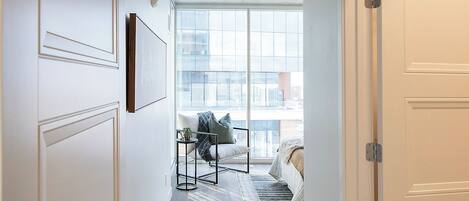 A hallway with a bed, a chair, a small table, and a window with a city view.