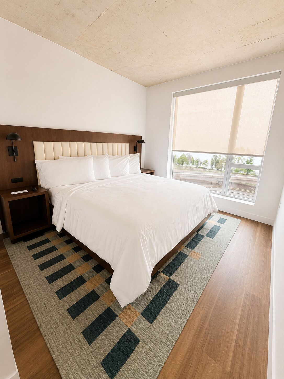 Un dormitorio con una cama grande, una mesita de noche, una lámpara de pared, una ventana con vistas y una alfombra estampada.