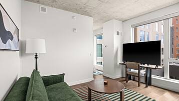 Una sala de estar moderna con un sofá verde, una lámpara, un televisor de pantalla plana, una mesita y vistas a un paisaje urbano a través de ventanas amplias.
