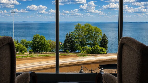 Quarto com uma grande janela com vista para a água, árvores e uma estrada.