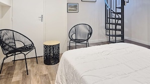 Quarto com cama, duas cadeiras, uma mesinha, escada em espiral e duas fotos emolduradas na parede.