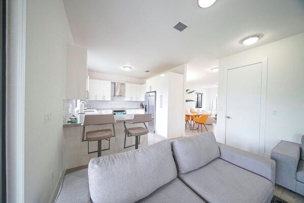 A modern living room with a grey sofa, a dining area with a table and chairs, and a kitchen with white cabinets and a refrigerator.