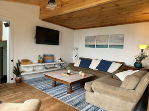 A living room with a wooden ceiling, a flat-screen TV, a sofa with blue and white cushions, a coffee table, and a potted plant.