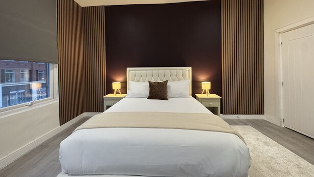 A modern bedroom with a large bed, two bedside tables with lamps, a dark accent wall, and a window with a view of buildings. - Hammersmith Haven (London)
