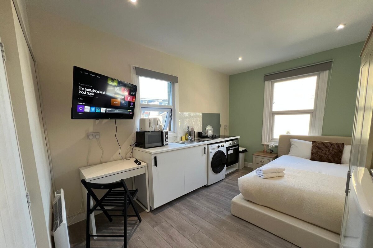 A compact hotel room with a kitchenette, a bed, a desk, and a TV mounted on the wall.