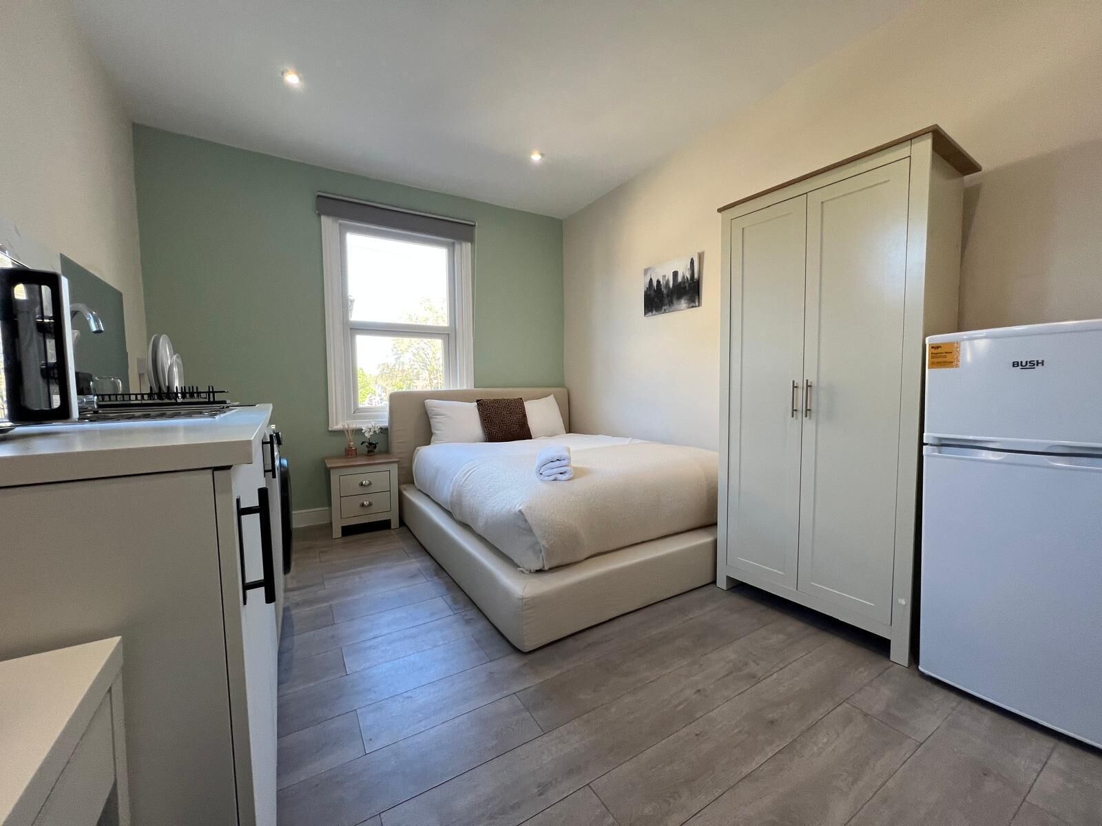 A bedroom with a bed, a desk with a computer, a wardrobe, and a refrigerator.