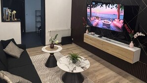 Moderni olohuone, jossa on suuri litteänäyttöinen televisio, musta sohva, kaksi sohvapöytää ja puulattia.