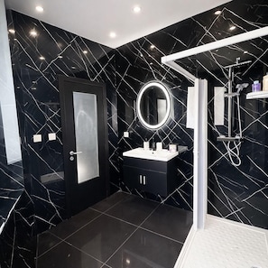 A modern bathroom with a black marble wall, a white sink, a round mirror, and a shower area.