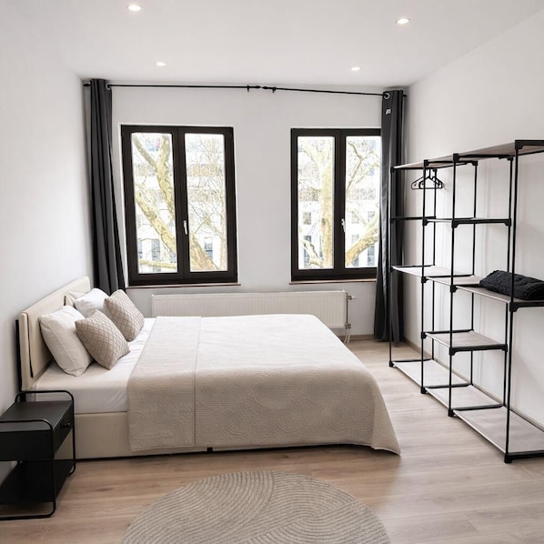 A modern bedroom with a bed, a black metal wardrobe, and two windows with a city view.