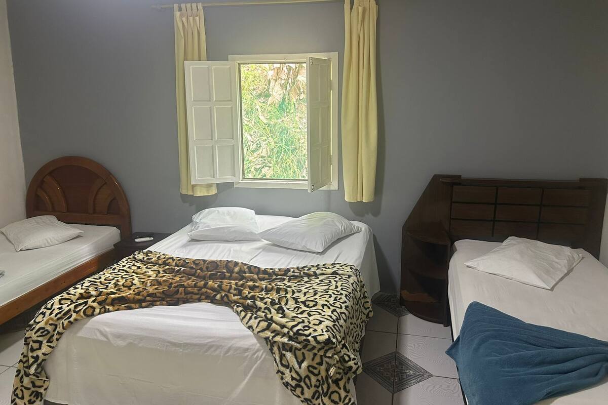 A room with two beds, a window with curtains, and a view of greenery outside.