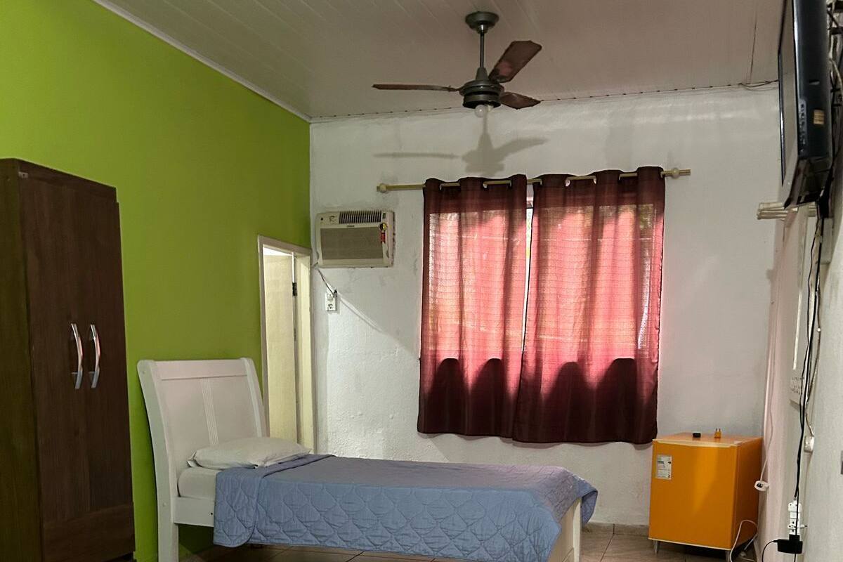A room with two beds, a ceiling fan, a window with curtains, a wardrobe, and a small orange cabinet.