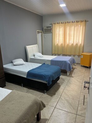 A hotel room with two beds, a small table, a cabinet, and a window with curtains. - pousada do pc (Bom Jesus do Itabapoana)