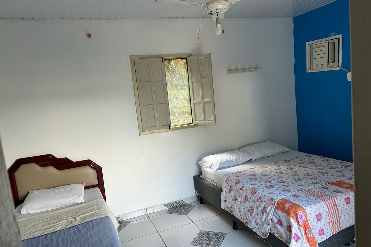 A bedroom with two beds, a ceiling fan, a window with shutters, and a wall-mounted air conditioning unit.